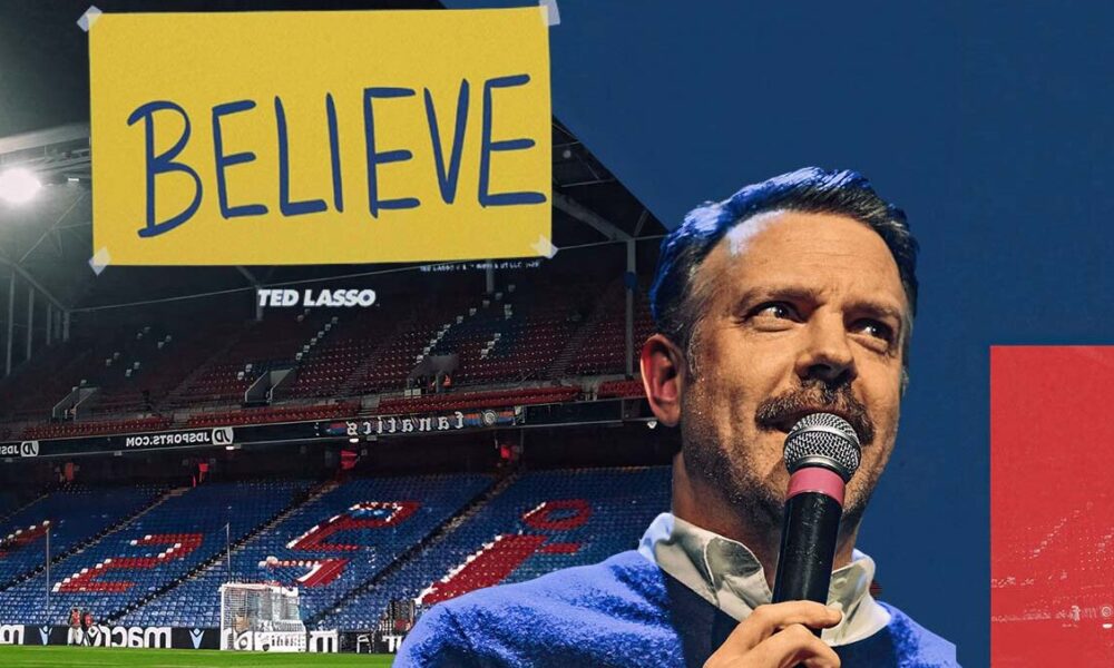 Ted Lasso speaking into a microphone with a 'Believe' banner in a soccer stadium, encapsulating the show's inspirational theme.