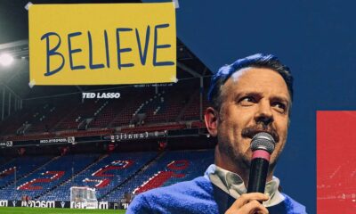 Ted Lasso speaking into a microphone with a 'Believe' banner in a soccer stadium, encapsulating the show's inspirational theme.