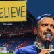 Ted Lasso speaking into a microphone with a 'Believe' banner in a soccer stadium, encapsulating the show's inspirational theme.