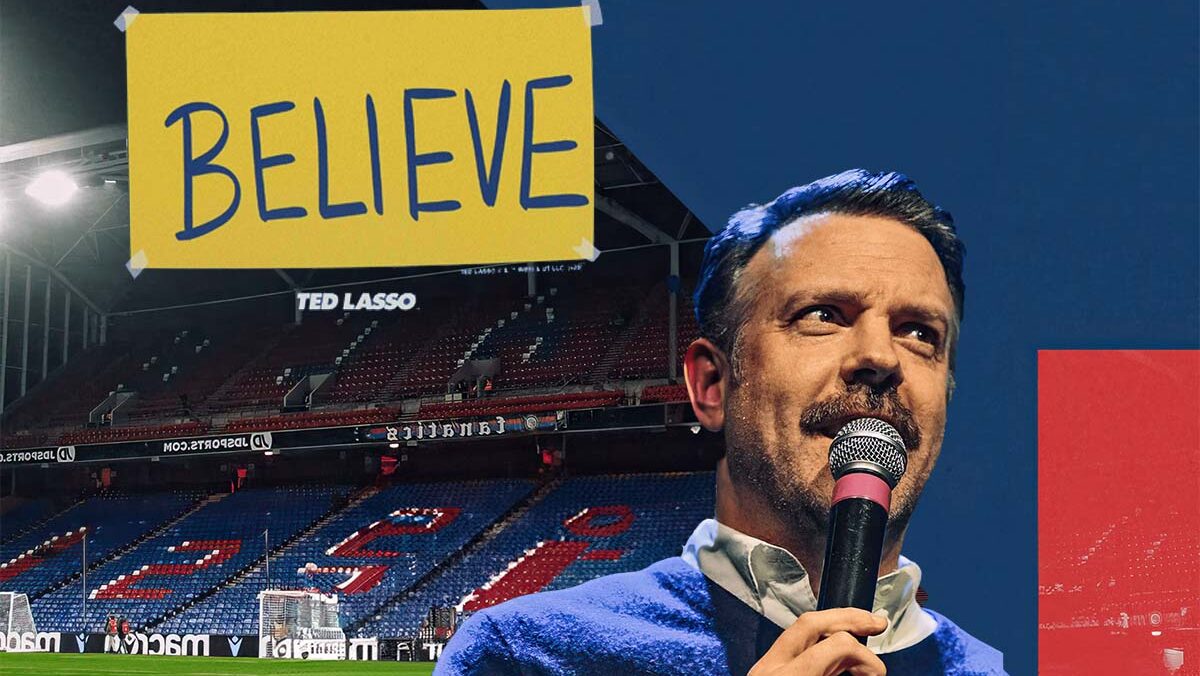 Ted Lasso speaking into a microphone with a 'Believe' banner in a soccer stadium, encapsulating the show's inspirational theme.